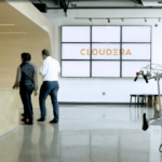 Cloudera office with guard behind wooden desk on left, bikes in standing bike rack on the right, and several people passing a large Cloudera logo in the foreground.